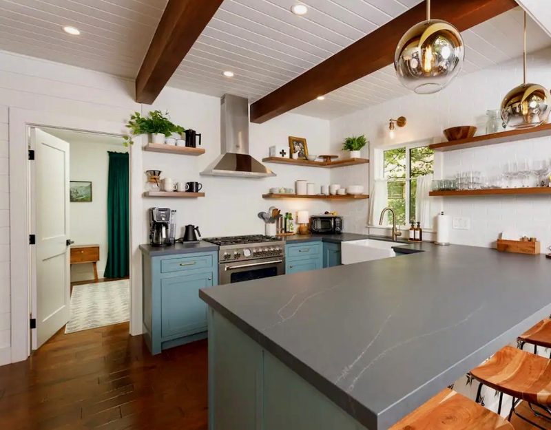 Bright kitchen with blue cabinets, open wood shelves and a gray matte quartz peninsula with wood stools